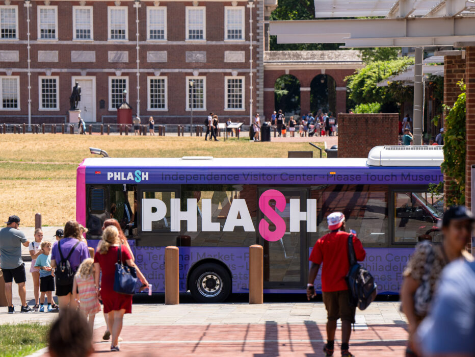 Philadelphia’s PHLASH bus is parked near Independence Mall as visitors walk along the sidewalk, with historic buildings and open green space visible in the background.