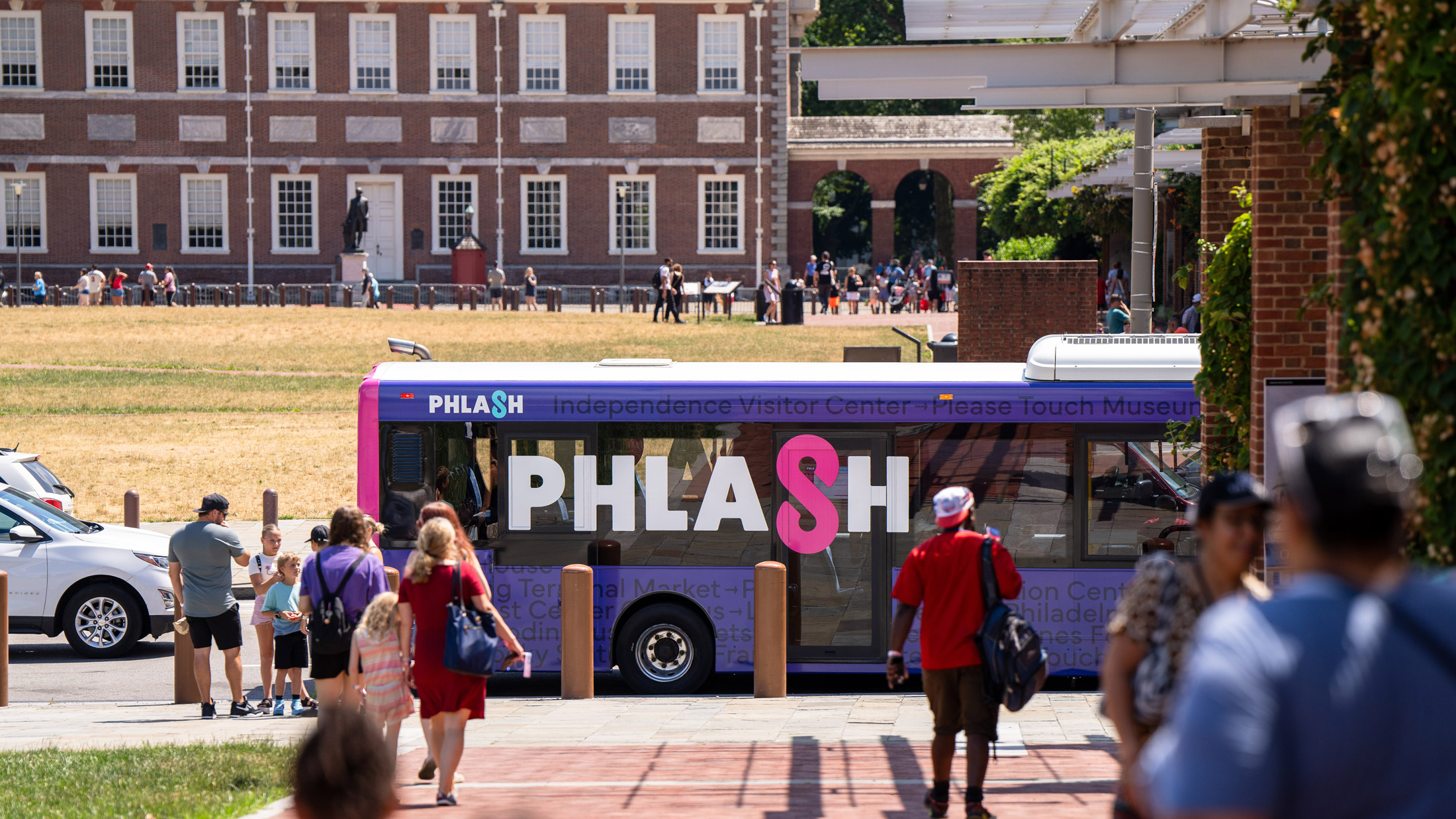 Philadelphia’s PHLASH bus is parked near Independence Mall as visitors walk along the sidewalk, with historic buildings and open green space visible in the background.