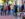 Group of young adults walking and chatting along a brick sidewalk in Phoenixville, Pennsylvania, passing small boutique storefronts with clothing displays outside while other people sit and socialize nearby.