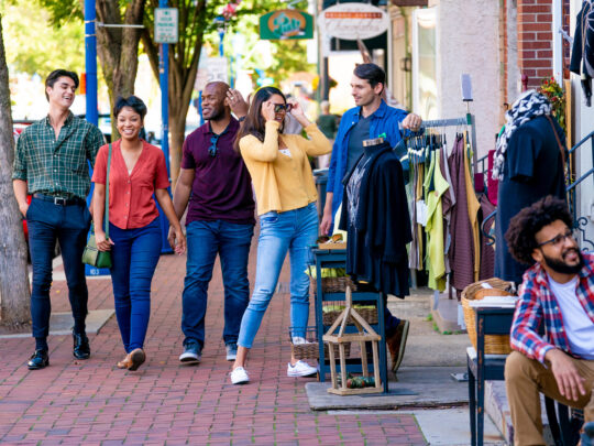 Un grupo de jóvenes camina y charla por una acera de ladrillo en Phoenixville, Pensilvania, pasando junto a pequeños escaparates de boutiques con ropa expuesta afuera, mientras otras personas se sientan y charlan cerca de allí.