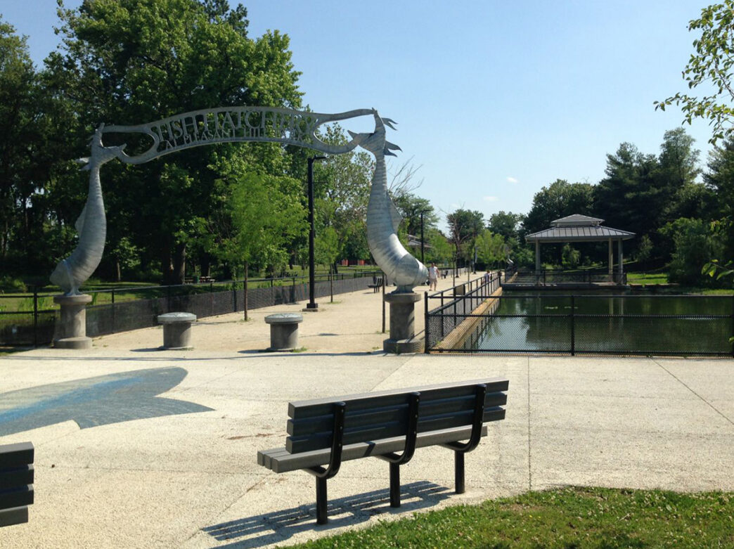 A decorative metal archway stands along a paved path beside a pond and pavilion in a leafy park setting.