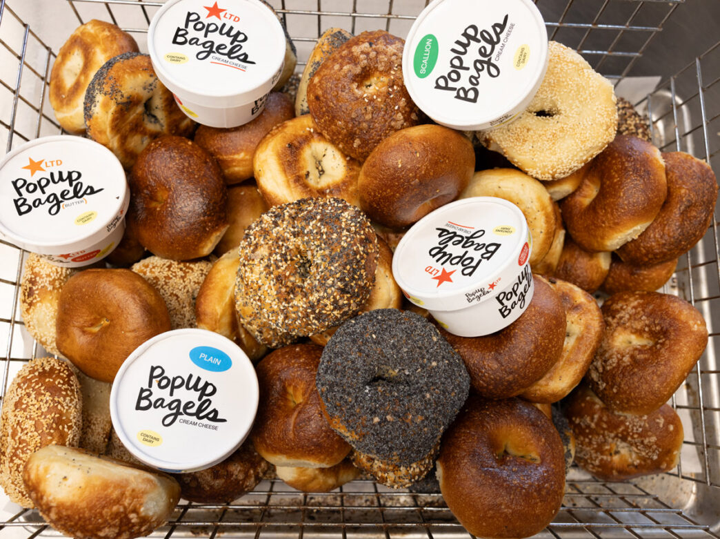 Un panier métallique rempli d'un assortiment de bagels fraîchement cuits - nature, au sésame, à tout faire et aux graines de pavot - surmonté de plusieurs contenants de fromage frais PopUp Bagels.
