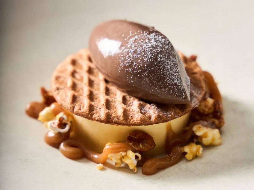 Artfully plated dessert at Provenance in Philadelphia featuring a round custard topped with a crisp wafer and a quenelle of chocolate cream, surrounded by caramel sauce, popcorn, and nuts.