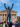 Rocky statue stands in front of the Philadelphia Museum of Art as a crowd of children and families celebrate Rocky Day, tossing their hands up while colorful confetti falls against a bright blue sky.