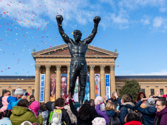 La estatua de Rocky se alza frente al Philadelphia Museum of Art mientras una multitud de niños y familias celebra el Día de Rocky, levantando los brazos mientras cae un confeti de colores bajo un cielo azul brillante.