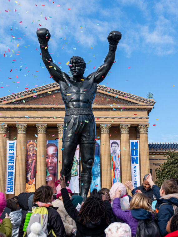 Rocky statue stands in front of the Philadelphia Museum of Art as a crowd of children and families celebrate Rocky Day, tossing their hands up while colorful confetti falls against a bright blue sky.