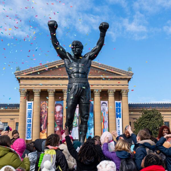 Rocky statue stands in front of the Philadelphia Museum of Art as a crowd of children and families celebrate Rocky Day, tossing their hands up while colorful confetti falls against a bright blue sky.
