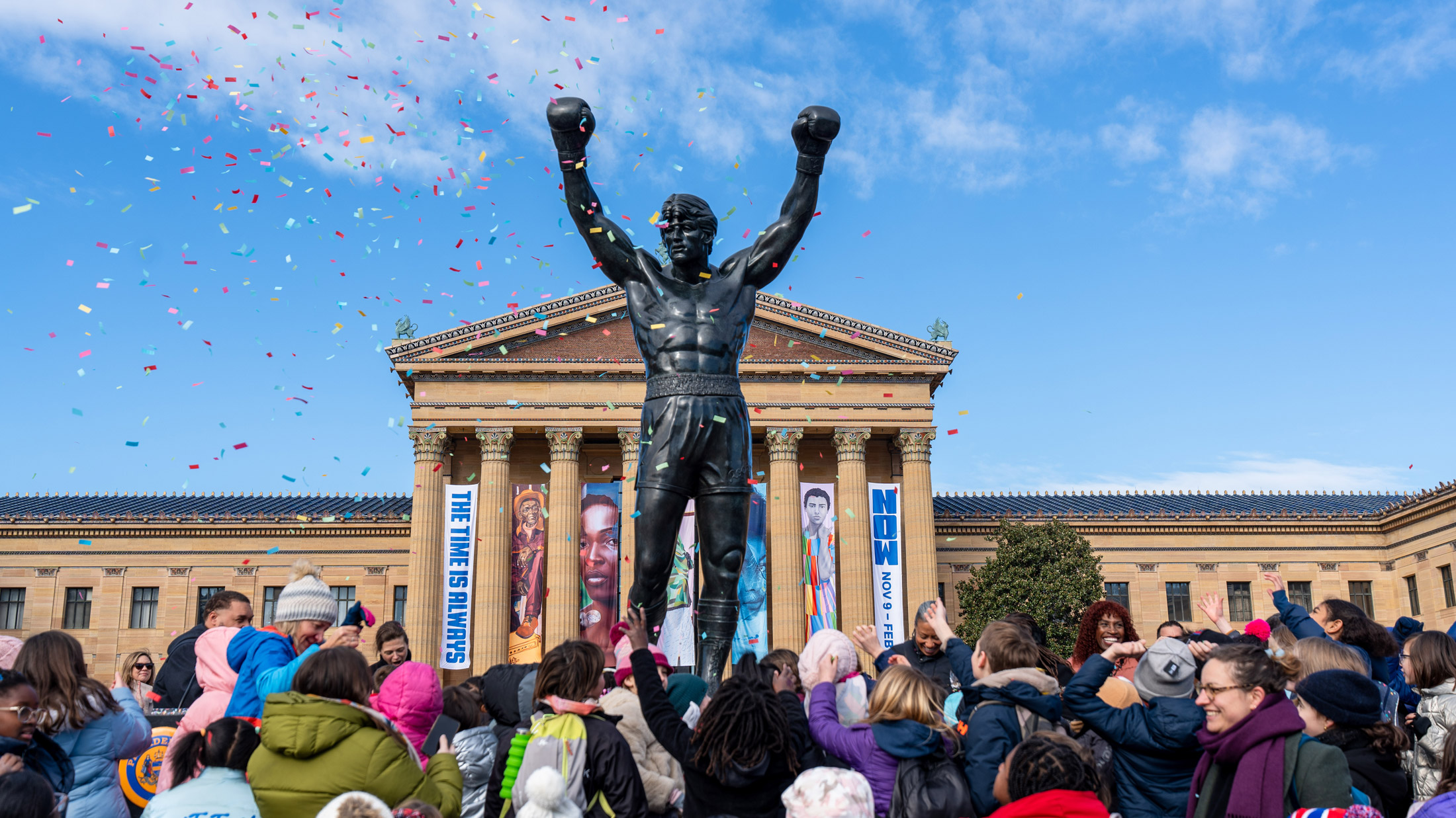 Rocky statue stands in front of the Philadelphia Museum of Art as a crowd of children and families celebrate Rocky Day, tossing their hands up while colorful confetti falls against a bright blue sky.