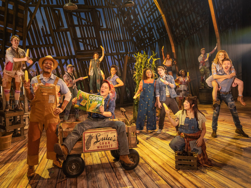 The cast of Shucked performs onstage in a barn set, dressed in colorful farm attire and holding ears of corn, with lively expressions and playful poses.