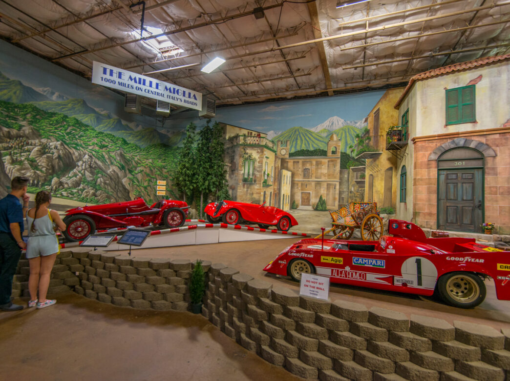 Visitors view vintage red race cars on display at the Simeone Foundation Automotive Museum in Philadelphia, set against a painted backdrop of an Italian village and countryside.