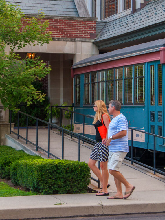 A couple walks past a colorful vintage trolley outside Hotel Fiesole in Skippack, Pennsylvania, surrounded by landscaped greenery and a brick building.