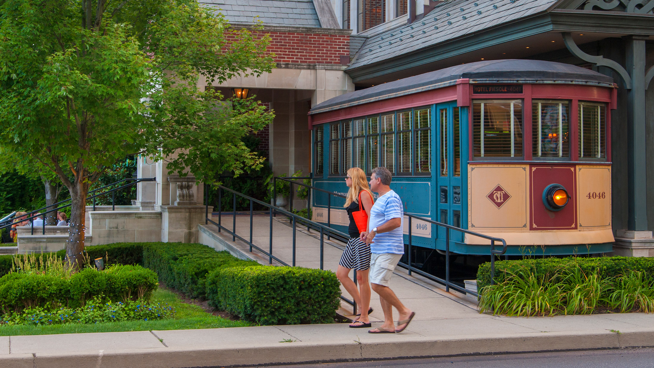 A couple walks past a colorful vintage trolley outside Hotel Fiesole in Skippack, Pennsylvania, surrounded by landscaped greenery and a brick building.