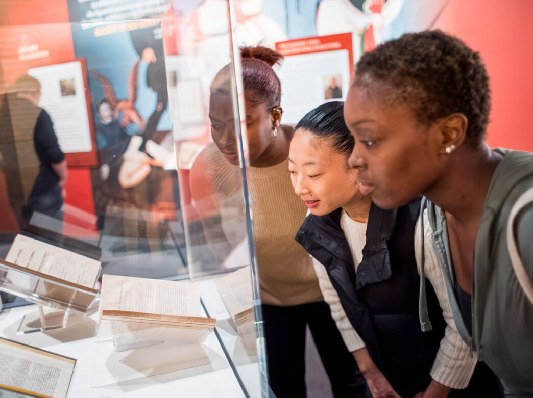Os visitantes se inclinam para examinar documentos históricos exibidos em uma caixa de vidro no Museum of the American Revolution na Filadélfia, com painéis interpretativos e exposições ao fundo.
