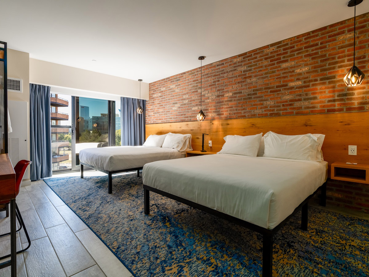 Guest room at The Maj Hotel in Philadelphia featuring two beds, a brick accent wall with pendant lighting, a patterned rug and a window overlooking the city.