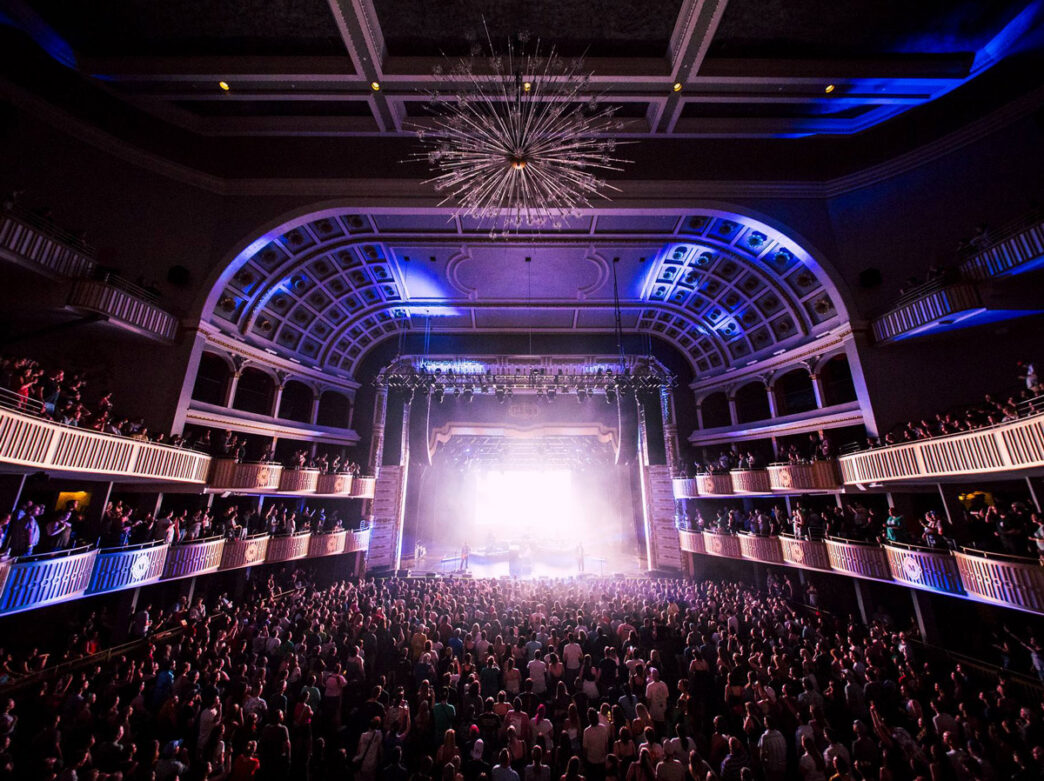 Una multitud abarrota el interior del Met de Filadelfia para asistir a un concierto, mientras las brillantes luces del escenario iluminan los ornamentados balcones del teatro y su gran candelabro.