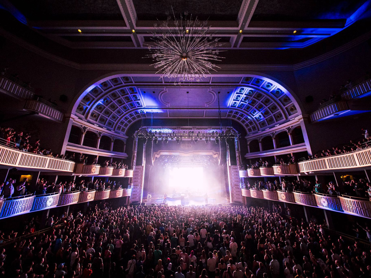 A packed crowd watches a concert inside The Met Philadelphia, with bright stage lights illuminating the theater’s ornate balconies and large chandelier.