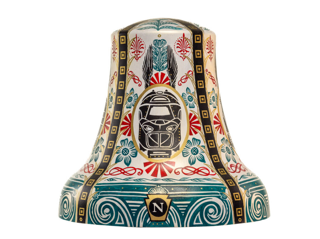 Bell sculpture titled “The Ringing Railroad” from Philadelphia250’s Bells Across PA, featuring a stylized train centered within an ornate, symmetrical design. The cream-colored surface is decorated with teal, red, and black patterns, including floral motifs and rail-like vertical bands, with a circular emblem containing an “N” for north near the base, evoking transit and connectivity in University City.