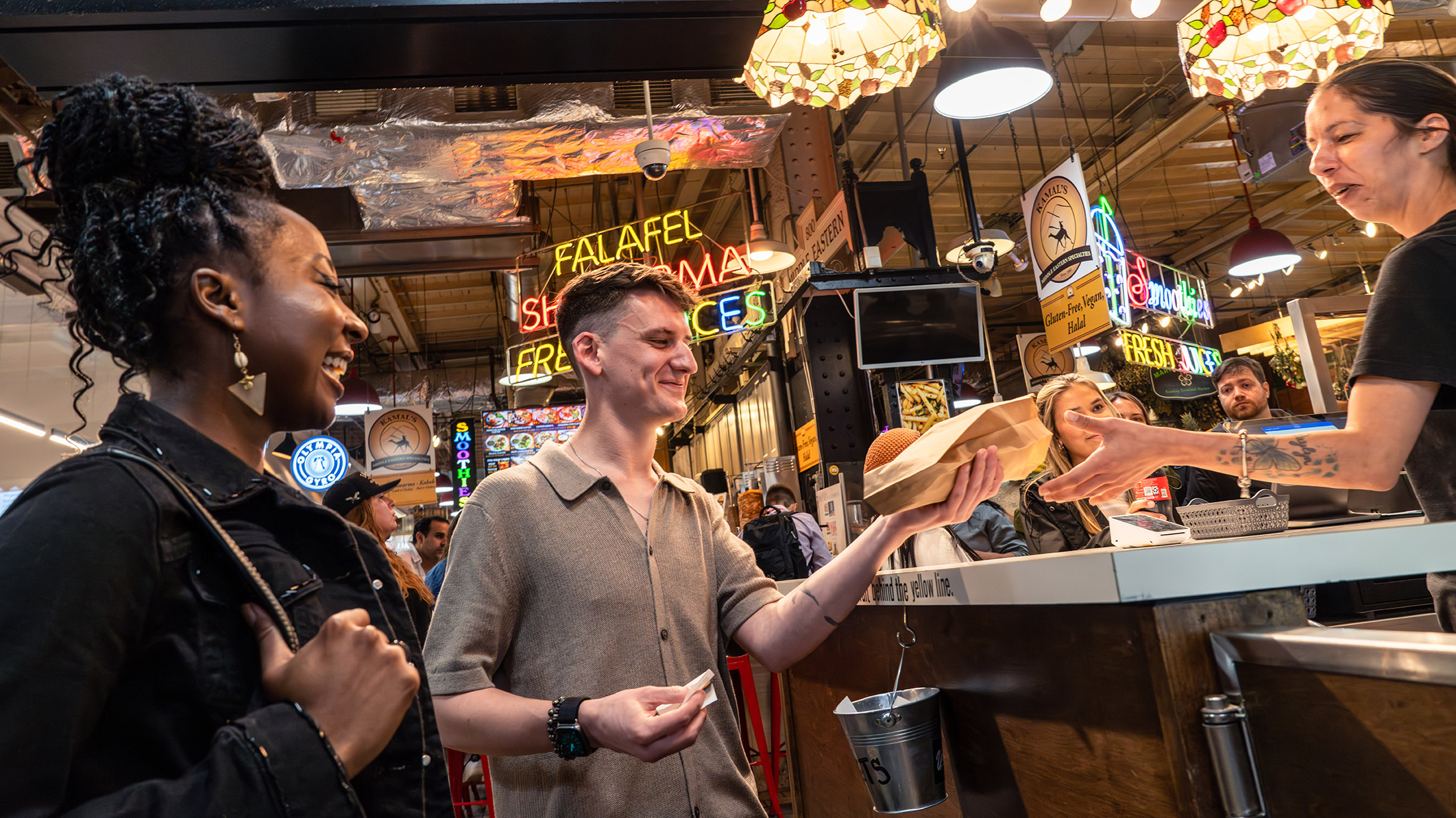 A man and a woman receiving a cheesesteak from a vendor at Reading Terminal Market. Patrons sit in the background beneath stained glass overhead lights