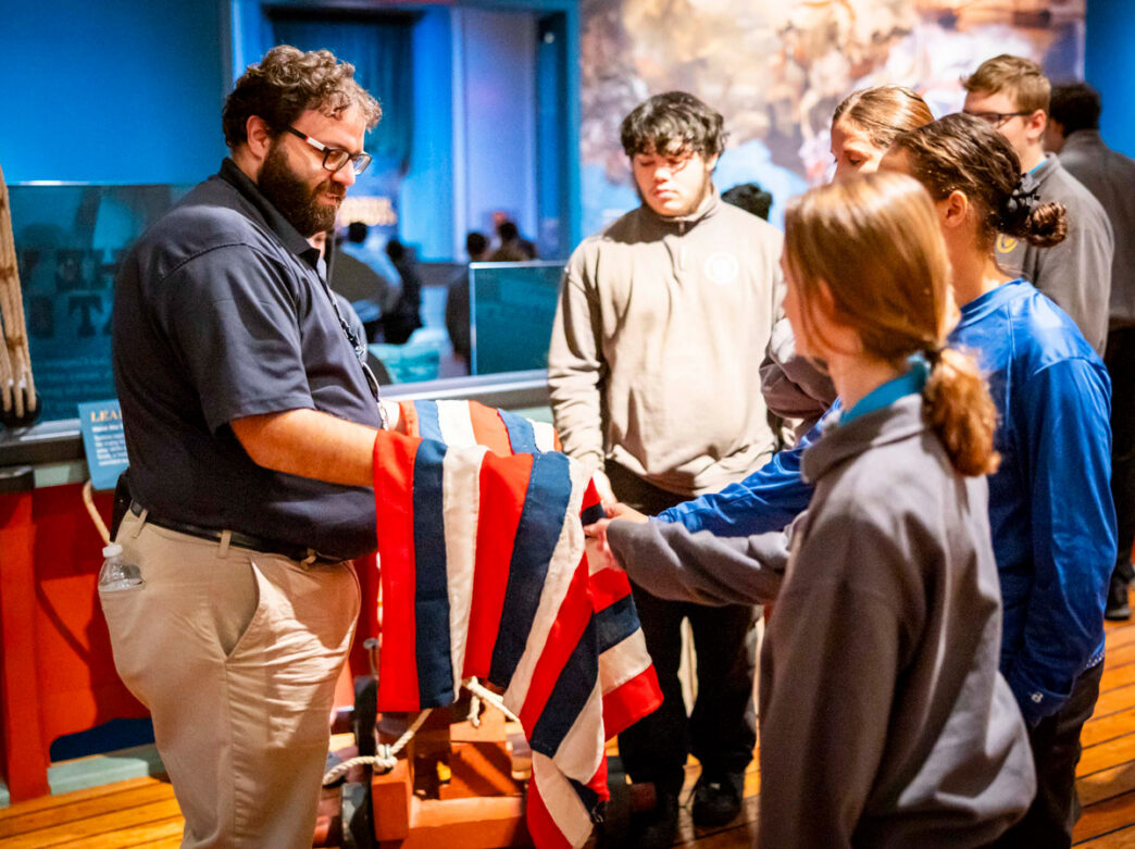 Young visitors listen to a guide and touch a historic flag as part of Unpacking the Sea Chest at the Museum of the American Revolution in Philadelphia.