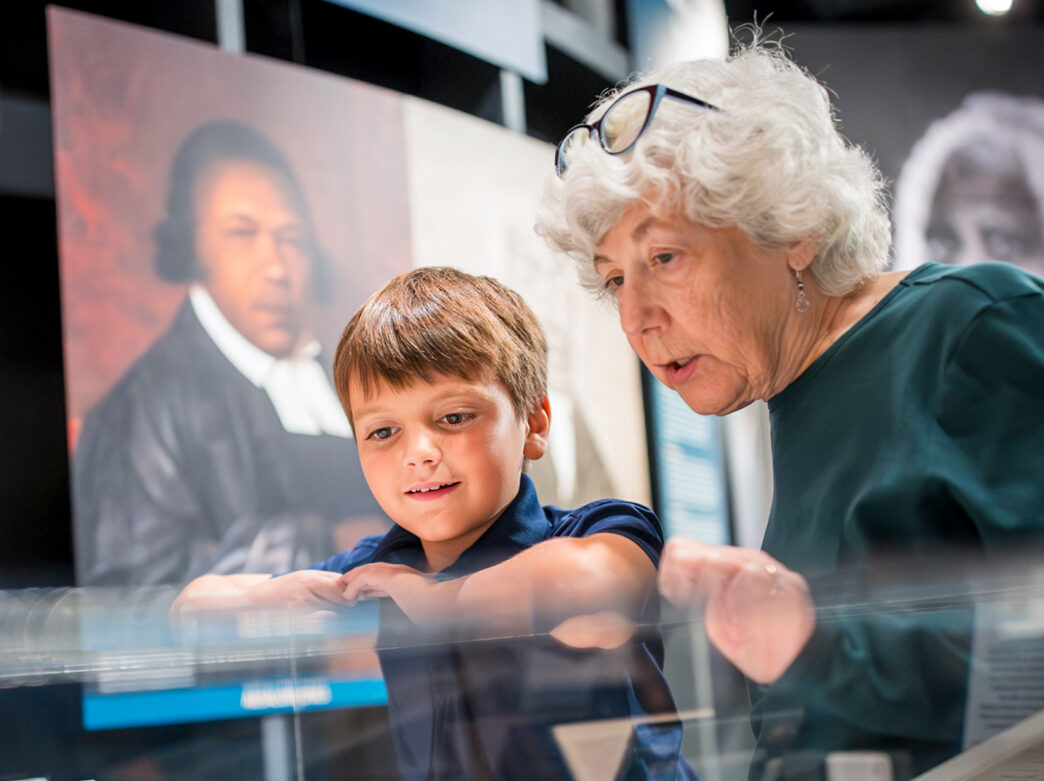 Two people look closely at artifacts inside a display case at the Museum of the American Revolution in Philadelphia, with historical portraits visible in the background.