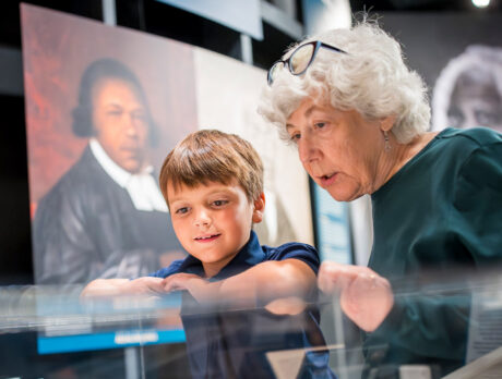 Two people look closely at artifacts inside a display case at the Museum of the American Revolution in Philadelphia, with historical portraits visible in the background.