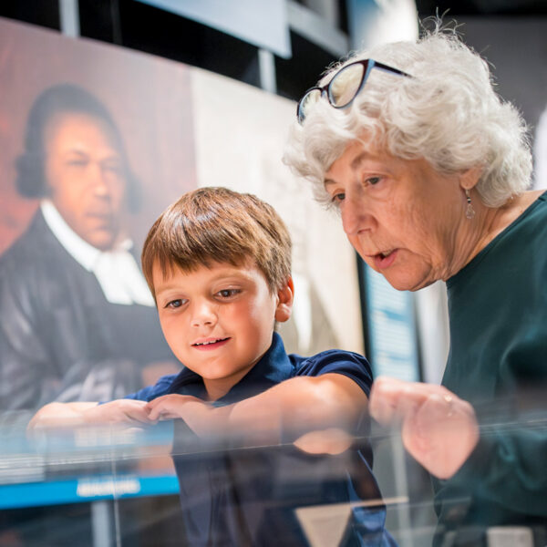 Two people look closely at artifacts inside a display case at the Museum of the American Revolution in Philadelphia, with historical portraits visible in the background.