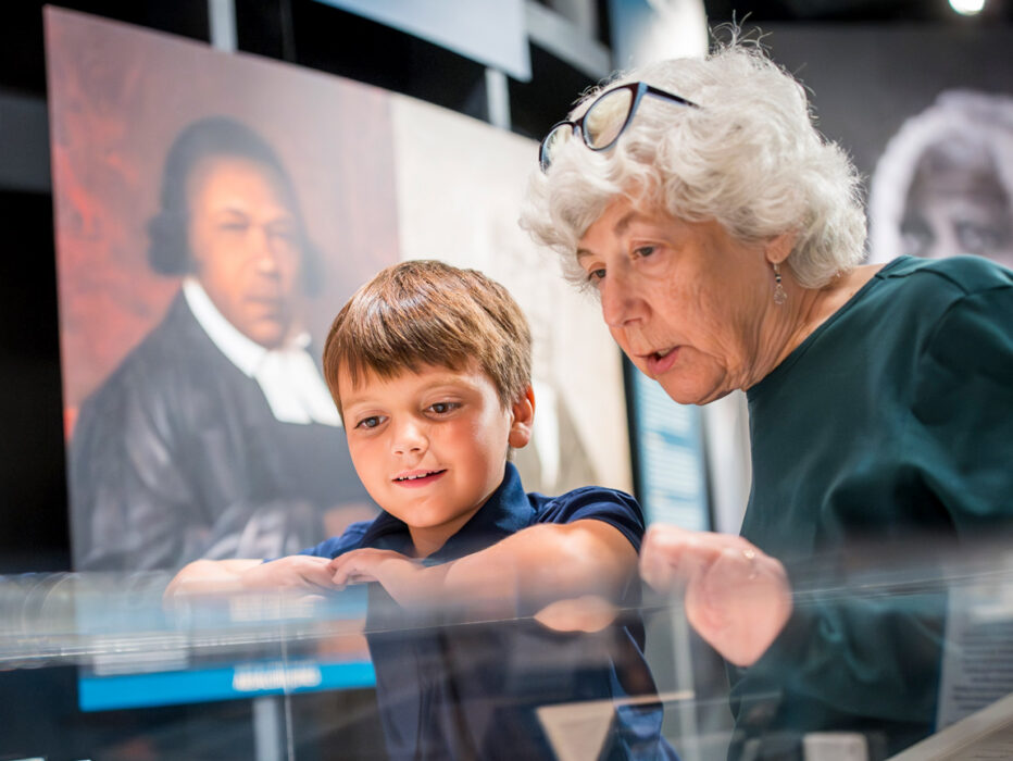 Dos personas observan con atención los objetos expuestos en una vitrina del Museum of the American Revolution de Filadelfia, con retratos históricos visibles al fondo.