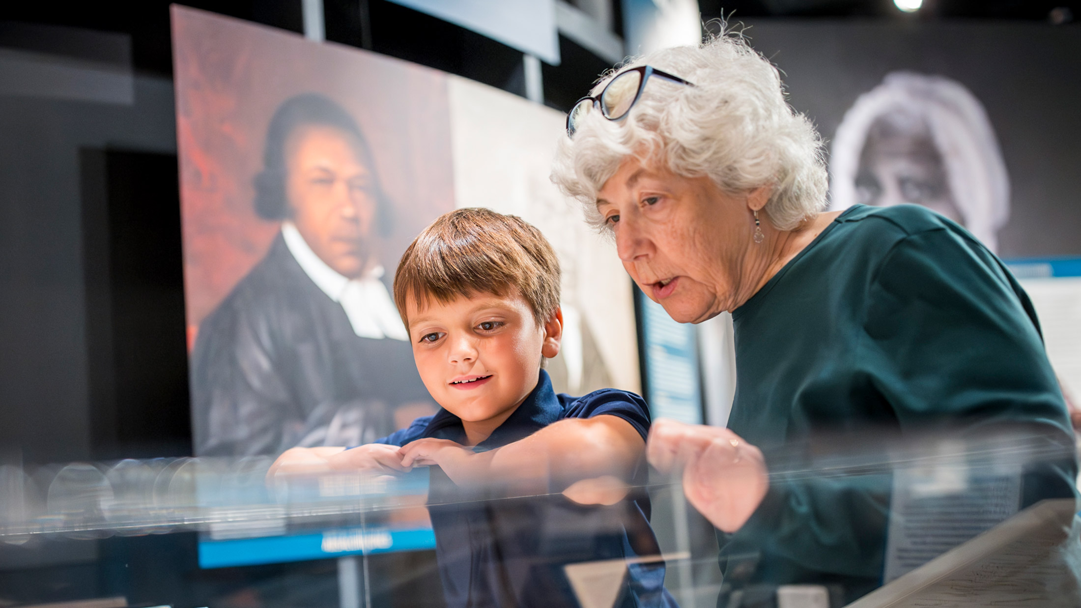 Two people look closely at artifacts inside a display case at the Museum of the American Revolution in Philadelphia, with historical portraits visible in the background.