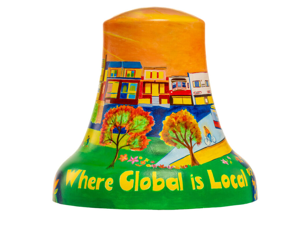 Bell sculpture titled “Where Global is Local” from Philadelphia250’s Bells Across PA, featuring a vibrant neighborhood streetscape. Colorful rowhomes line the background beneath a warm orange sky, with trees, flowers, and a cyclist in the foreground. The phrase “Where Global is Local” appears in large yellow lettering along the green base, highlighting Olney’s diverse community.