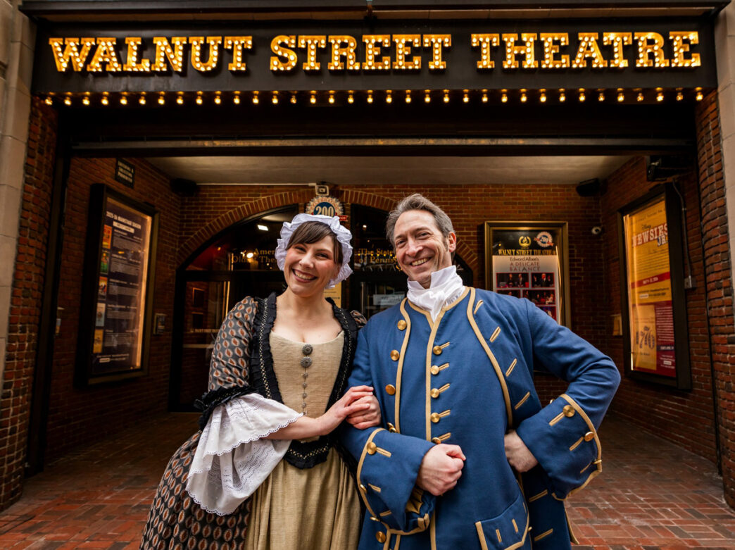 Two actors in 18th-century costume from 1776: The Musical pose smiling outside the Walnut Street Theatre, beneath its illuminated marquee.