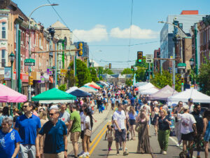 Mensenmassa's wandelen over een afgesloten Girard Avenue tijdens het Girard Avenue Street Fest in de wijk Brewerytown in Philadelphia, omzoomd door kleurrijke verkooptenten en historische rijtjeshuizen op een zonnige dag.