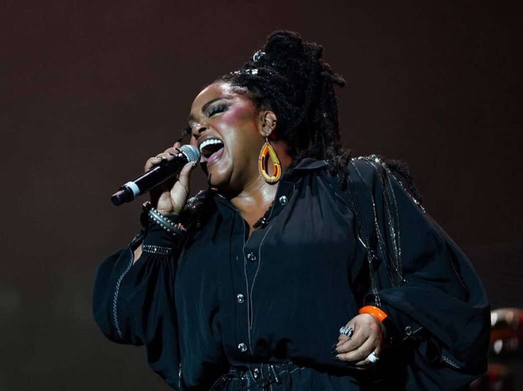 Jill Scott sings into a microphone while performing on stage. Her eyes are closed.