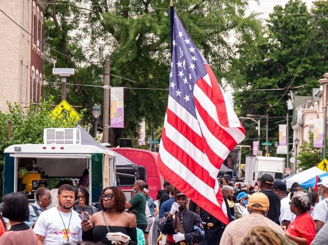 A gentleman in military dress attire carries a large American flag through the crowd at the Philadelphia Juneteenth Festival in Germantown.