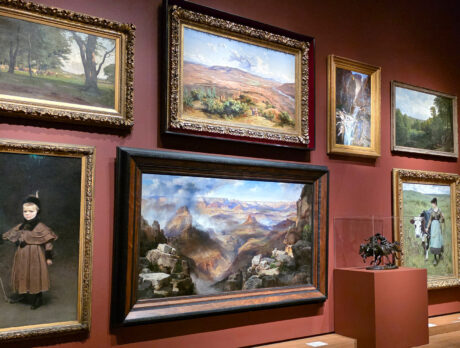 Gallery wall in the “A Nation of Artists” exhibit in Philadelphia displaying a salon-style arrangement of framed landscape and portrait paintings, with a visitor viewing the artwork.