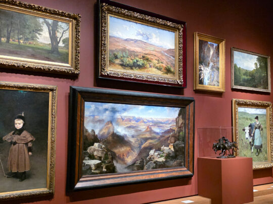 Gallery wall in the “A Nation of Artists” exhibit in Philadelphia displaying a salon-style arrangement of framed landscape and portrait paintings, with a visitor viewing the artwork.