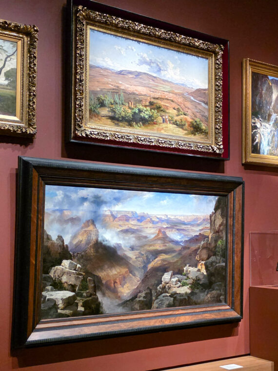 Gallery wall in the “A Nation of Artists” exhibit in Philadelphia displaying a salon-style arrangement of framed landscape and portrait paintings, with a visitor viewing the artwork.