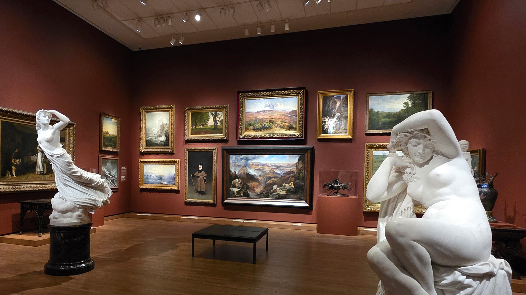 Gallery view inside Philadelphia’s A Nation of Artists exhibit featuring red walls lined with framed landscape and figure paintings, with two white marble sculptures of women displayed prominently in the foreground and center of the room.
