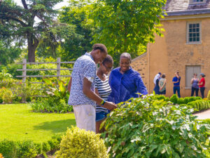 A family smiles and examines plants in a lush garden outside a historic stone building in Philadelphia's Germantown neighborhood on a sunny day.