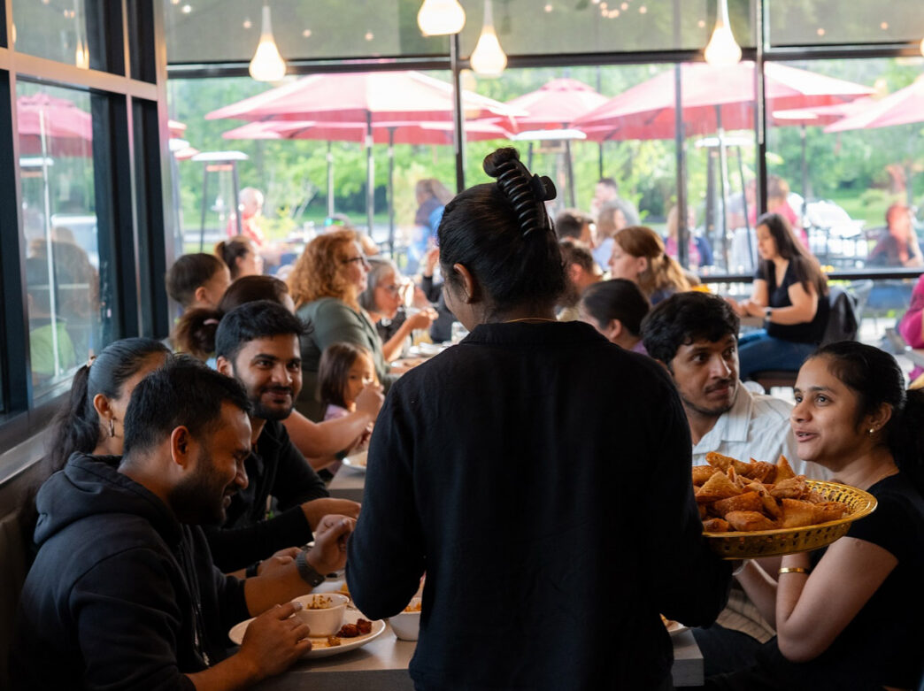 A waitress at Amma's South Indian Cuisine in Philadelphia holds a plate of samosas while speaking to a group dining at a table.