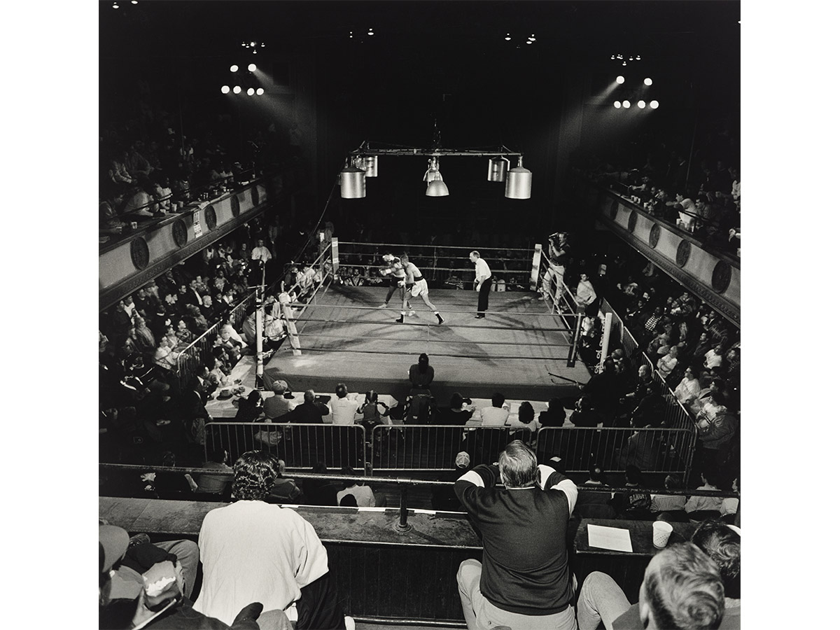 A dramatic black and white aerial photograph of a boxing match in progress at Philadelphia's legendary Blue Horizon venue, with a packed crowd of spectators filling the intimate balconied hall under bright ring lights.