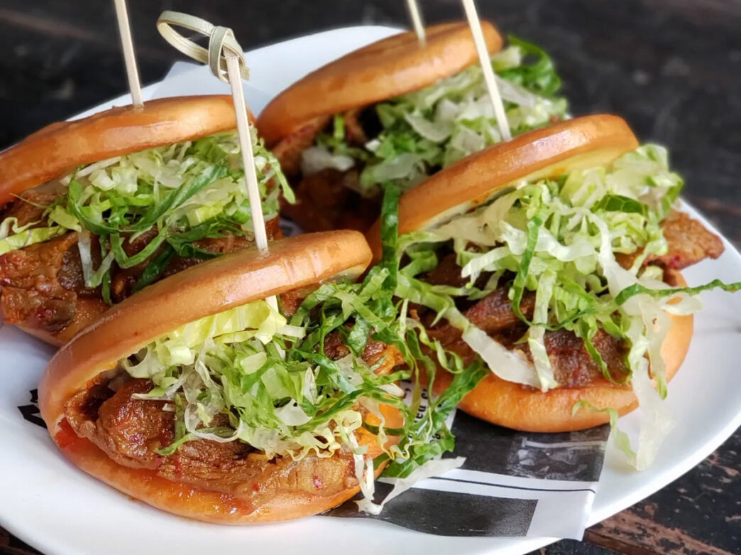 Close-up of four Korean-style pork belly slider buns from Buk Chon in Philadelphia, topped with shredded lettuce and secured with wooden picks on a white plate.