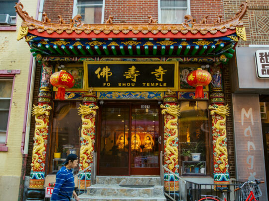 La entrada, ricamente decorada, del templo Fo Shou, situado en el Chinatown de Filadelfia, presenta elementos arquitectónicos tradicionales chinos, como aleros curvos, dragones dorados, linternas rojas y pilares con elaboradas tallas de motivos tradicionales.
