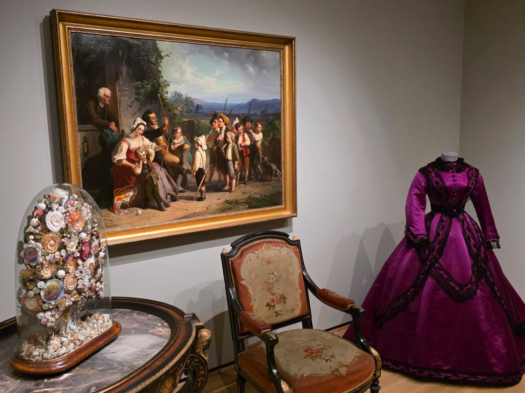 Galerievertoning van de tentoonstelling A Nation of Artists in Philadelphia met een ingelijst 19e-eeuws schilderij, een Victoriaanse magenta jurk op een paspop en antiek meubilair waaronder een stoel en een decoratieve glazen schikking.