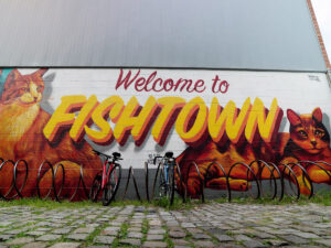 Two bikes parked at a spiral bike rack in front of a colorful "Welcome to Fishtown" street mural featuring large painted cats, on a cobblestone street in Philadelphia's Fishtown neighborhood.