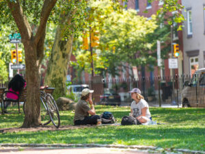 Two people sit on the grass in the shade of a tree at Fitler Square park in Philadelphia, chatting on a sunny day with a bicycle, park bench, and brick rowhouses visible nearby.