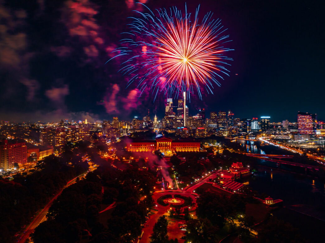 Uma vista aérea dramática de fogos de artifício vermelhos, brancos e azuis explodindo sobre o Philadelphia Museum of Art e a Benjamin Franklin Parkway no dia 4 de julho, com o horizonte da cidade iluminado ao fundo.
