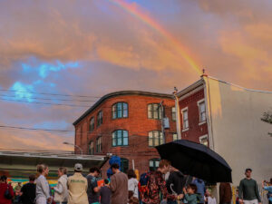 People gather outside under a partly-cloudy sky with a rainbow in the Francisville neighborhood of Philadelphia.