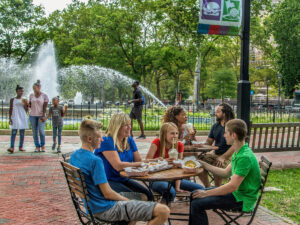Groups of visitors enjoy food and drinks at outdoor tables in Franklin Square, one of Philadelphia's original five squares, with the park's iconic fountain and lush trees visible in the background.