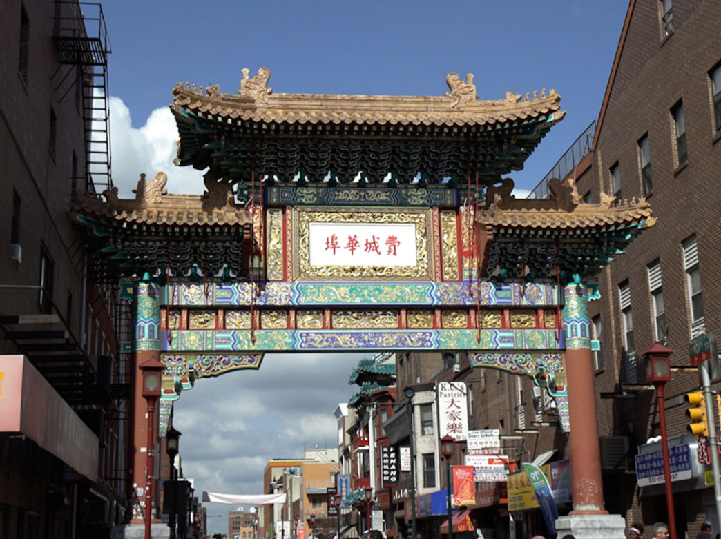 The ornate Friendship Gate marks the entrance to Philadelphia's Chinatown neighborhood, spanning 10th Street with intricate gold, red, and teal traditional Chinese architectural details against a partly cloudy sky.