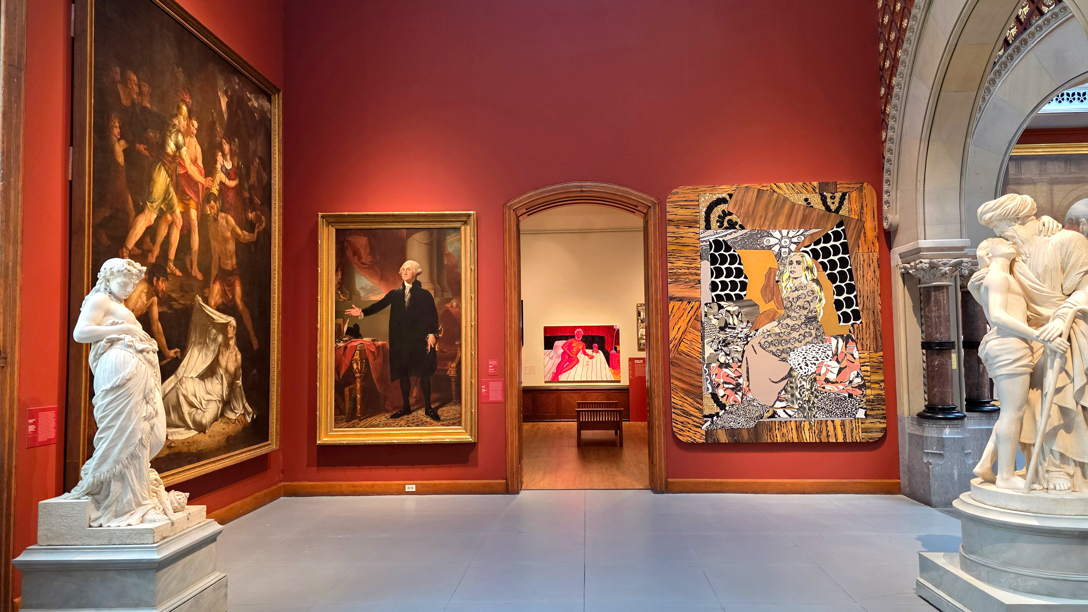 Gallery view in Philadelphia’s A Nation of Artists exhibit with deep red walls, featuring large historic and contemporary paintings—including a portrait of George Washington—flanked by white marble sculptures and an arched doorway leading to another room.